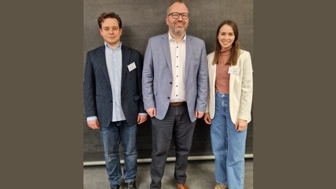 Drei erwachsene Personen stehen nebeneinander vor einer dunklen Wand, frontal zur Kamera. Alle tragen legere bis formelle Kleidung mit Namensschildern; links ein Mann im Sakko, in der Mitte ein Mann im hellblauen Sakko, rechts eine Frau im hellen Blazer.