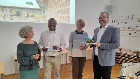 Vier Personen stehen in einem hellen Seminarraum nebeneinander und lächeln in die Kamera. Im Hintergrund sind Tische, Wandplakate und eine große Projektion zu sehen.