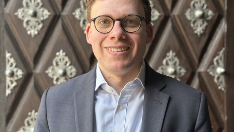 Mann mit blonden Haaren und Brille steht lächelnd vor einer verzierten Holzwand/Tür. Er trägt ein hellgraues Jackett und ein hellblaues Hemd