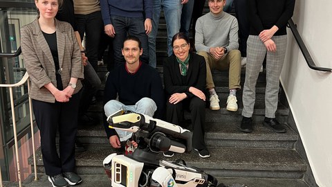 Menschen stehen und sitzen auf einer Treppe. Vor ihnen steht ein Roboter mit vier Beinen und länglichem Körper.