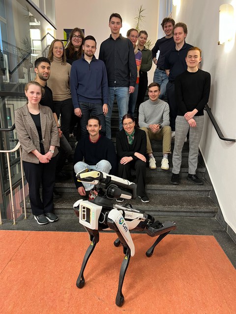 Menschen stehen und sitzen auf einer Treppe. Vor ihnen steht ein Roboter mit vier Beinen und länglichem Körper.