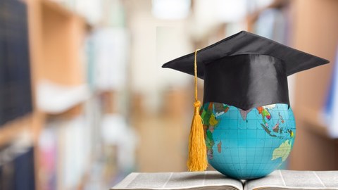 Bild von einem kleinen Globus, der auf einem Buch liegt und eine Graduiertenmütze aufhat in einer Bibliothek.
