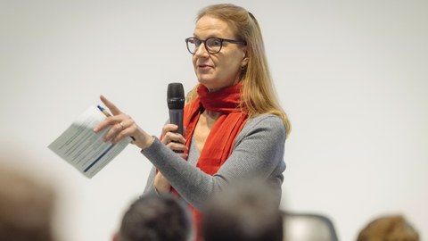 Prof. Marianne Kneuer spricht mit einem Mikrofon in der Hand vor einer Gruppe Menschen.