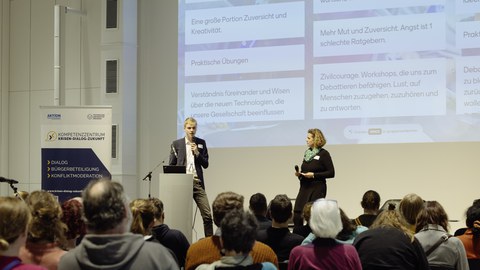 Eine Gruppe Menschen sitzt vor einer Bühne. Auf dieser stehen ein Mann und eine Frau mit Mikrofonen und sprechen.