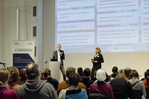 Eine Gruppe Menschen sitzt vor einer Bühne. Auf dieser stehen ein Mann und eine Frau mit Mikrofonen und sprechen.