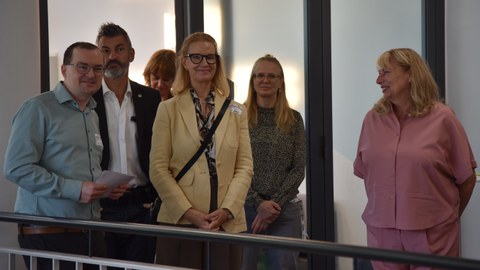 Eine Gruppe Personen vor einer Glastür, im Vordergrund ein Geländer.