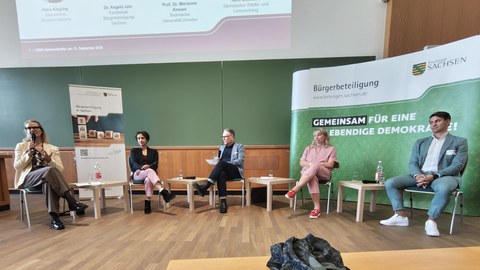 Eine Podiumsdiskussion mit fünf Teilnehmern in einem Hörsaal. Im Hintergrund ein Aufsteller "Bürgerbeteiligung in Sachsen".