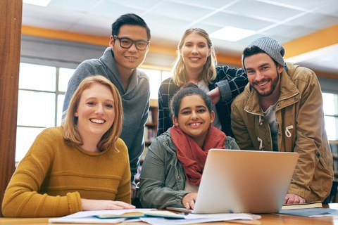 Foto einer Gruppe von 5 Studenten, die gemeinsam in der Bibliothek zusammen lernen und fröhlich in die Kamera lächeln