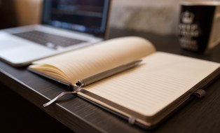 Arbeitsplatz mit Laptop, offenem Notizbuch mit Stift und einer Tasse im Hintergrund.