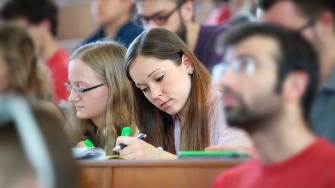 Das Foto zeigt einen Hörsaal voller Studierender. Der Fokus liegt dabei auf zwei Studentinnen, die mit grünen Textmarkern etwas in ihren Arbeitsmaterialien markieren.