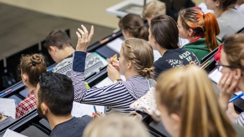 Foto von Studierenden in einer Vorlesung in einem Hörsaal. Eine davon meldet sich.