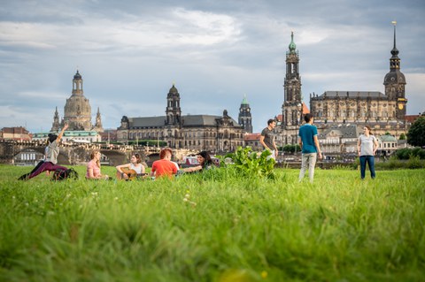 Gruppe von Studenten sitzt und steht plaudernd an der Elbe. Eine spielt Gitarre und zwei machen Yoga.