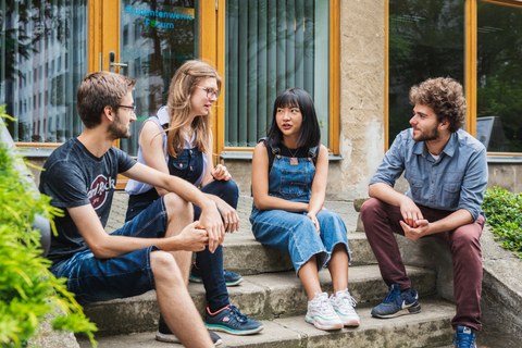 Foto: Im Freien sitzen vier Studierende auf einer Treppe und unterhalten sich.