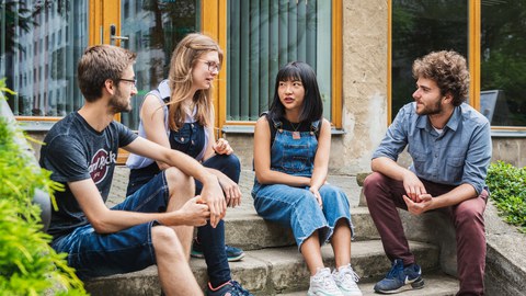 Foto: Im Freien sitzen vier Studierende auf einer Treppe und unterhalten sich.
