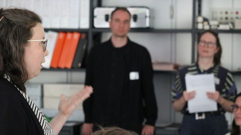Drei Personen bei einem Vortrag. Links eine Frau im Profil, die gerade spricht. In der Mitte zwei stehende Personen, die zuhören. 