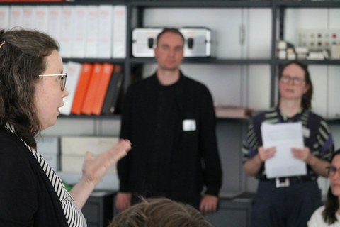Drei Personen bei einem Vortrag. Links eine Frau im Profil, die gerade spricht. In der Mitte zwei stehende Personen, die zuhören. 