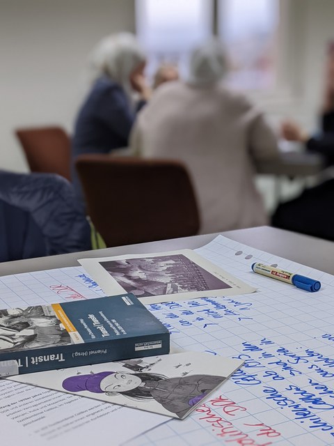 Ein Tisch mit Büchern, Postkarten und Papier im Vordergrund. Verschwommen im Hintergrund Menschen an einem anderen Tisch.