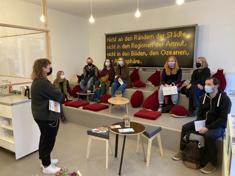 Die Erstsemester:innen des Fachbereichs Kunstgeschichte im Kunsthaus Dresden. Sie sitzen auf einer treppenförmigen Sitzgelegenheit mit roten Kissen unter einer großen Anzeigetafel mit dem Schriftzug "Nicht an den Rändern der Städte, nicht in den Regionen der Armut, nicht in den Böden, den Ozeanen, der Atmosphäre". Vor Ihnen steht eine Mitarbeiterin des Kunsthauses. Der Raum ist hell in grau und weiß gehalten, es gibt Beistelltische und eine Art Bar. 