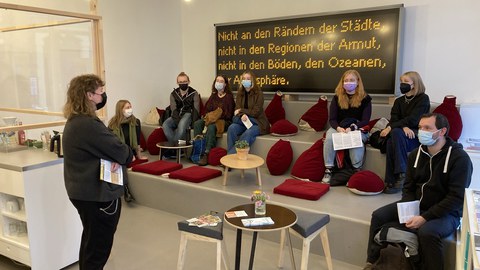 Die Erstsemester:innen des Fachbereichs Kunstgeschichte im Kunsthaus Dresden. Sie sitzen auf einer treppenförmigen Sitzgelegenheit mit roten Kissen unter einer großen Anzeigetafel mit dem Schriftzug "Nicht an den Rändern der Städte, nicht in den Regionen der Armut, nicht in den Böden, den Ozeanen, der Atmosphäre". Vor Ihnen steht eine Mitarbeiterin des Kunsthauses. Der Raum ist hell in grau und weiß gehalten, es gibt Beistelltische und eine Art Bar. 