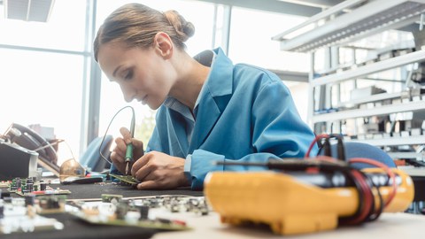 Foto einer jungen Frau, die in einer Fabrik ein Bauteil lötet.