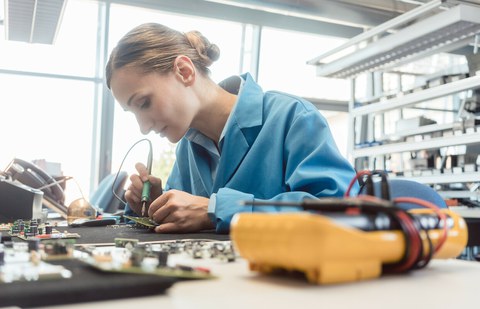 Foto einer jungen Frau, die in einer Fabrik ein Bauteil lötet.