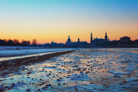 Das Foto zeigt die Elbwiesen im Winter. Die Wiese ist von einer Schicht aus Eis und Schnee überzogen. Im Hintergrund erkennt man die Silhouette der Dresdener Altstadt.