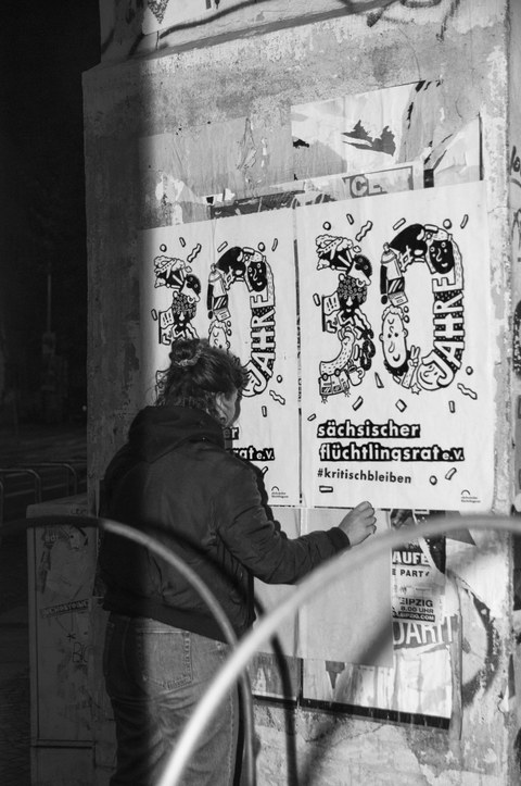 S/W-Foto: Eine Person klebt Plakate mit der Aufschrift "30 Jahre Sächsischer Flüchtlingsrat e.V., #kritischbleiben"