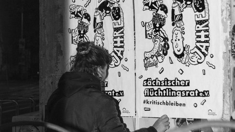 S/W-Foto: Eine Person klebt Plakate mit der Aufschrift "30 Jahre Sächsischer Flüchtlingsrat e.V., #kritischbleiben"