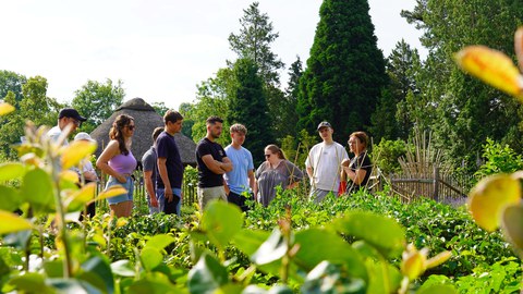 Ackercoachin und Dozentin Heidelinde Wutzler und eine Gruppe Student:innen in einem Garten. Im Vordergrund Pflanzen. 
