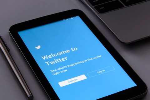 Ansicht des blauen Anmeldebildschirms der Twitter-App auf einem Handy, daneben liegt ein Stift bzw. ein Teil eines Laptops ist erkennbar.