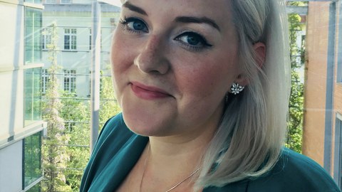 Porträtfoto von Anne-Marie Lachmund. Sie steht lächelnd im Halbprofil vor einer Glasscheibe.