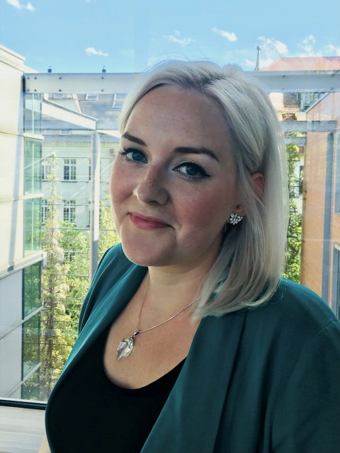 Porträtfoto von Anne-Marie Lachmund. Sie steht lächelnd im Halbprofil vor einer Glasscheibe.
