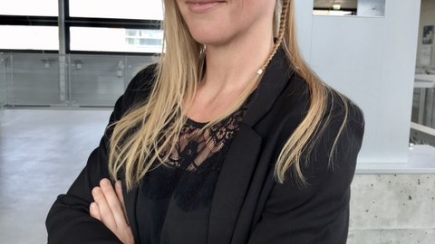 Porträfoto von Anna Rebecca Hoffmann: Sie steht im Halbprofil mit verschränkten Armen in einem großem hellen Raum und lächelt in die Kamera.