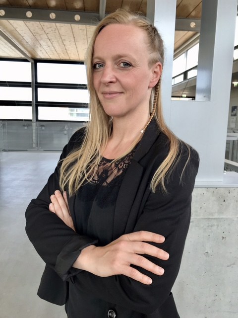 Porträfoto von Anna Rebecca Hoffmann: Sie steht im Halbprofil mit verschränkten Armen in einem großem hellen Raum und lächelt in die Kamera.
