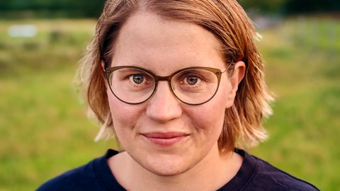 Porträtfoto von Anne Piezunka: Sie blickt frontal in die Kamera, lächelt dabei, trägt eine Brille und ein dunkles Oberteil. Sie steht auf einer Wiese, im Hintergrund sind unscharf Bäume zu erkennen.