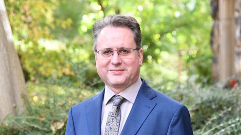 Porträtfoto von Jörg Ganzenmüller: Er steht im Anzug und mit Krawatte vor Bäumen und Büschen mit grünen Blättern. Er trägt eine Brille und lächelt in die Kamera.