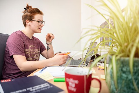 Eine Studentin sitzt an ihrem Schreibtisch, sie hat einen Kugelschreiber in der Hand. Vor ihr ein Laptop und ein Spiralblock, sie scheint sich mit jemandem per Videochat zu unterhalten. Im Vordergrund ein Buch, eine TUD-Tasse und Sachsengras.