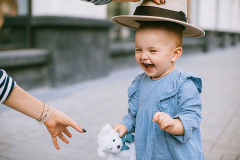 Auf dem Bild ist ein lachendes Kind zu sehen, welchem von einer erwachsenen Person ein Hut über den Kopf gehalten wird.