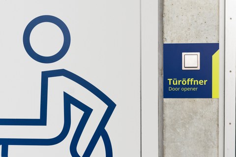 Das Bild ein Rollstuhlfahrer:innen-Piktogramm, daneben ist der Schalter zum Öffnen der Tür für Rollstuhlfahrende zu sehen. Es handelt sich um eine Aufnahme aus dem Hörsaalzentrum der TU Dresden..
