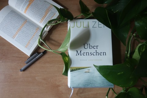 Eine Ausgabe von Juli Zehs "Über Menschen" liegt auf einem Tisch, umgeben von den Ranken einer Pflanze. Daneben ein aufgeschlagenes Buch, in dem Textpassagen markiert sind. 