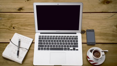 Das Bild zeigt einen von oben fotografierten Arbeitsplatz mit Laptop, Smartphone, Kaffee und aufgeschlagenem Notizbuch
