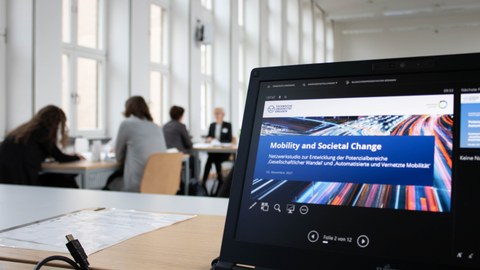 Laptopbildschirm, Ansicht einer Powerpointpräsentation. Startfolie mit dem Titel "Mobility and Societal Change". Im Hintergrund verschwommen Menschen. 