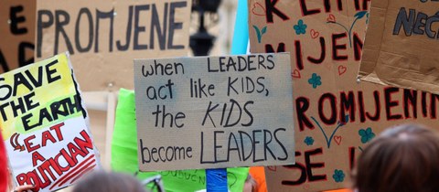 Zu sehen sind verschiedene beschriftete Pappschilder bei einer Demonstration. Eines der Schilder liest: When leaders act like kids the kids become leaders.