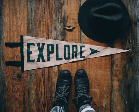 Zu sehen ist ein dreieckiges weißes Banner auf einem hölzernen Fußboden, bedruckt mit dem Wort Explore. Unter dem Banner sind ein paar Schuhe zu sehen, daneben ein Hut. 