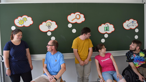 Gruppenbild vom QuaBIS Team vor einer Tafel. Über den Köpfen der Personen sind Sprechblasen zu sehen mit verschiedenen Symbolen. Zum Beispiel Fragezeichen, eine Glühbirne und ein Megafon.