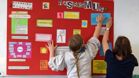 Zwei Frauen stehen mit dem Rücken zum Betrachter und strecken ihre Hände nach einem Plakat aus, auf dem der Begriff Rassismus erklärt wird.