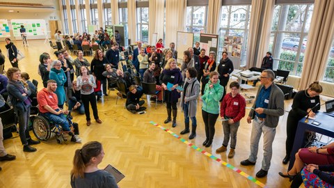 Das Foto zeigt viele Personen bei einer Inklusionsveranstaltung im Dülfersaal der TU Dresden. Sie stehen in einer großen Gruppe zusammen. Im Vordergrund stehen fünf Personen an einer Linie aus verschiedenfarbigen Kärtchen.