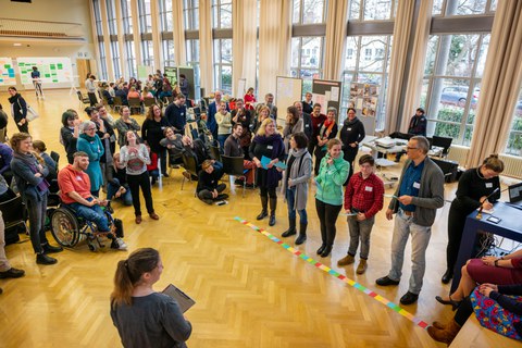 Das Foto zeigt viele Personen bei einer Inklusionsveranstaltung im Dülfersaal der TU Dresden. Sie stehen in einer großen Gruppe zusammen. Im Vordergrund stehen fünf Personen an einer Linie aus verschiedenfarbigen Kärtchen.