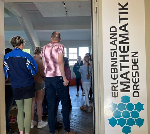 Auf dem Foto ist eine Gruppe von Personen von hinten zu sehen. Sie stehen in einer Eingangstür. Neben der Tür hängt ein Plakat mit der Aufschrift "Erlebnisland Mathematik Dresden"