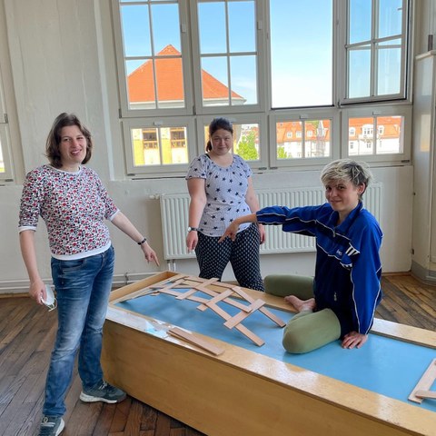 Auf dem Foto sind drei Personen zu sehen. Zwei stehen um einen rechteckigen Holzklotz und eine Person sitzt darauf. Alle drei zeigen mit der Hand auf eine kleine Brücke die auf dem Holzklotz aus Holzstäben gebaut ist. Alle lächeln fröhlich. 